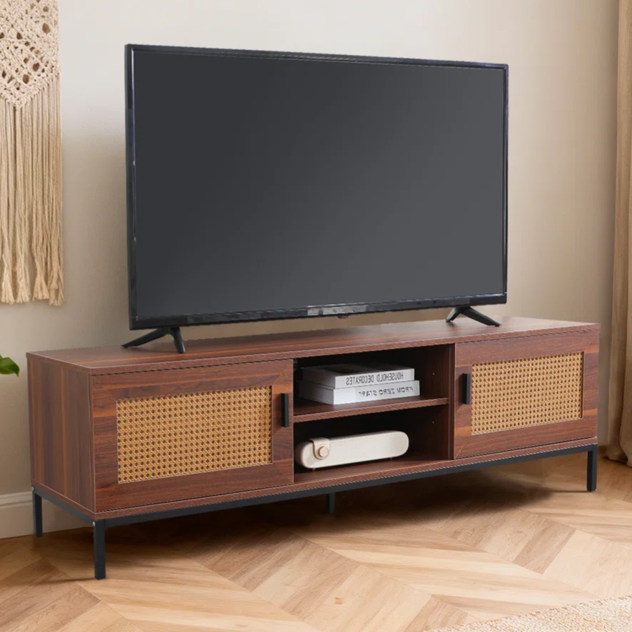 A flat-screen TV on a low wooden TV stand with cane accents is shown, accompanied by indoor plants, beige curtains, a macramé wall hanging, and a book.