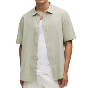 A person is wearing an open short-sleeve, light green button-up shirt over a white tank top, paired with light-colored pants.