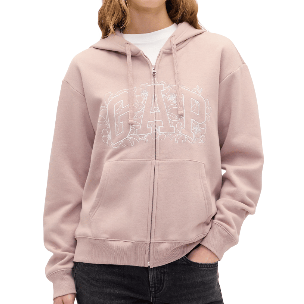 A person wearing a light pink GAP zip-up hoodie with white floral embroidery on the logo, paired with dark jeans.