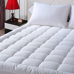 A white queen-sized mattress pad with a quilted pillow-top design displayed on a bed next to a nightstand with a red lamp.