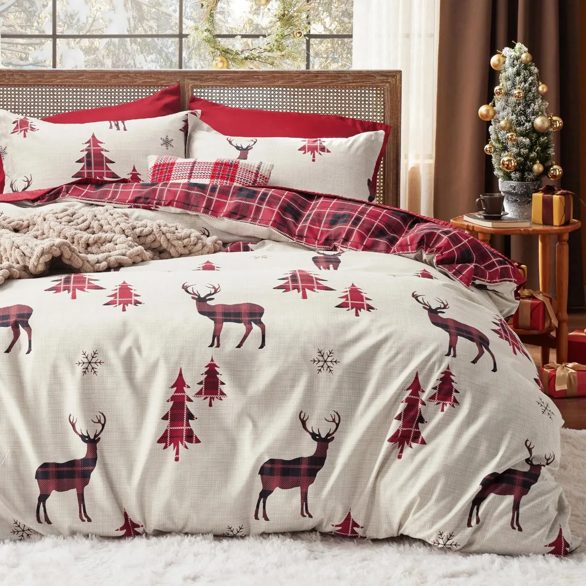 A cozy bedding set features a holiday theme with red and black plaid deer, red trees, and snowflakes, accented with a plaid throw blanket and decorative pillows. A small decorated Christmas tree and wrapped gifts sit on a side table.
