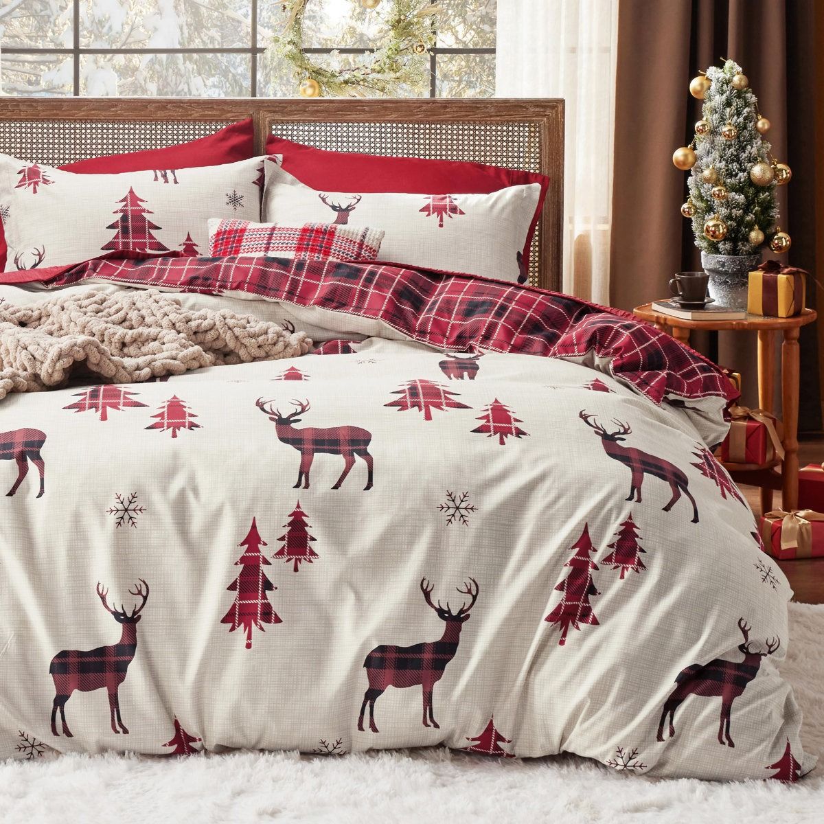 A cozy bedding set features a holiday theme with red and black plaid deer, red trees, and snowflakes, accented with a plaid throw blanket and decorative pillows. A small decorated Christmas tree and wrapped gifts sit on a side table.