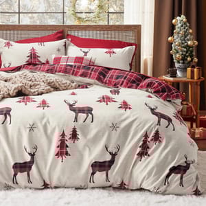 A cozy bedding set features a holiday theme with red and black plaid deer, red trees, and snowflakes, accented with a plaid throw blanket and decorative pillows. A small decorated Christmas tree and wrapped gifts sit on a side table.