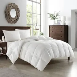 A bedroom features a white comforter on a bed with wooden headboard, two pillows, a nightstand, a decorative mirror, and a tall dresser with plants and decor. Large windows let in natural light.