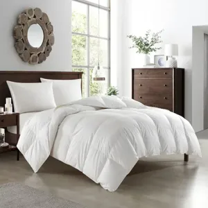 A bedroom features a white comforter on a bed with wooden headboard, two pillows, a nightstand, a decorative mirror, and a tall dresser with plants and decor. Large windows let in natural light.