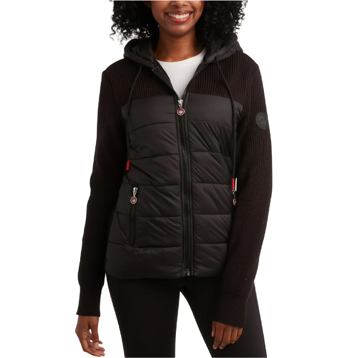 A woman is wearing a black zip-up jacket with a padded front and knitted sleeves. The jacket features a hood, zippered pockets, and a small logo patch on the sleeve.