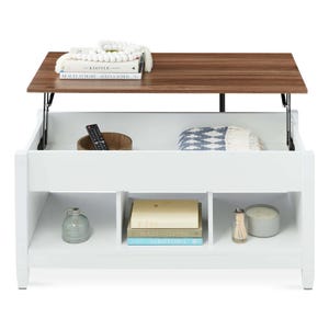 A white lift-top coffee table with storage compartments, featuring a wood top that raises, revealing space for a blanket and a remote. Books and decorative items are stored below.