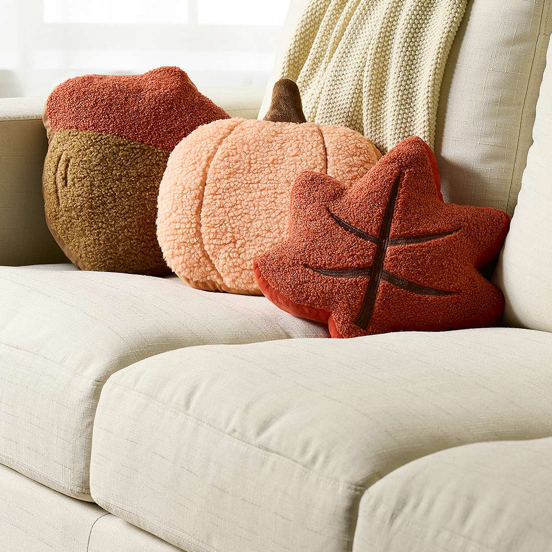 Three plush pillows shaped like fall-themed items—a rust-colored leaf, a tan acorn, and a round orange pumpkin—are arranged on a cream sofa.