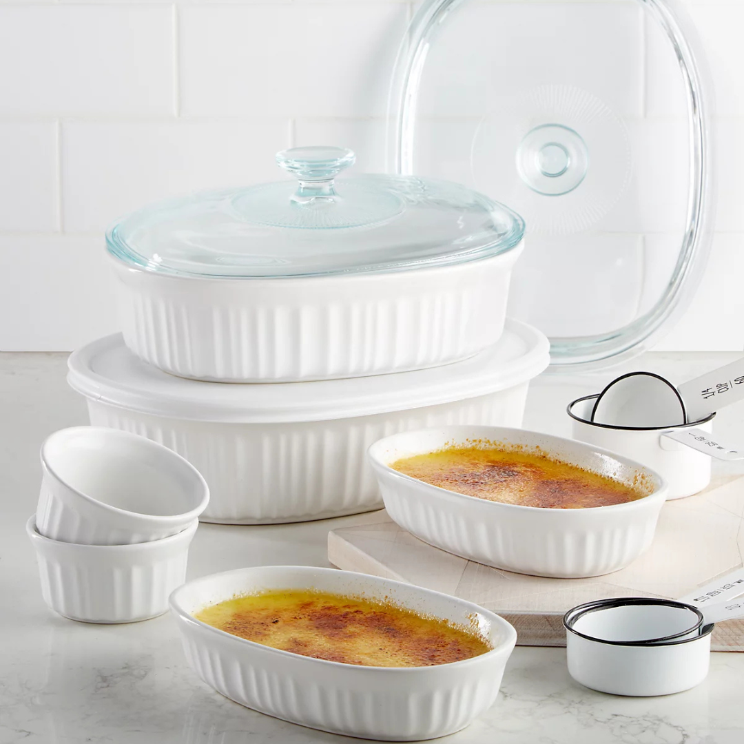 White ceramic bakeware set with ribbed design, includes various dishes, bowls, and a measuring cup set. Some dishes have glass lids, and creme brulee is shown in two of the dishes.