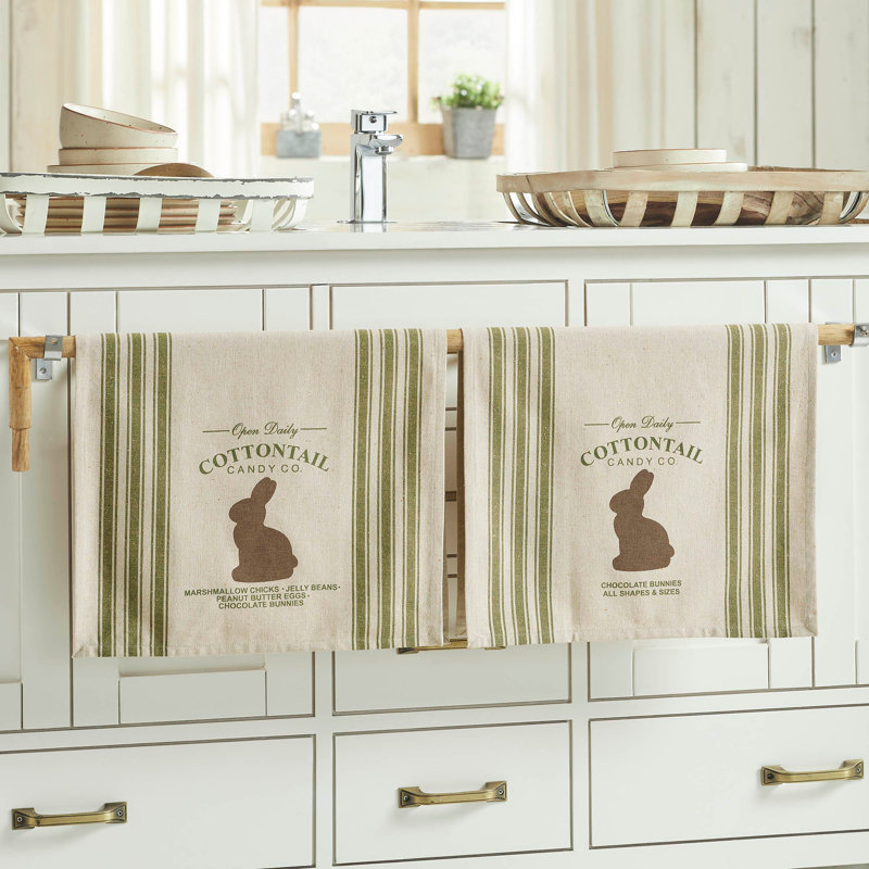 Two kitchen towels hang on a cabinet, featuring green stripes and a brown bunny with the text \“Cottontail Candy Co.\“ They list candy types like marshmallow chicks, jelly beans, and chocolate bunnies.