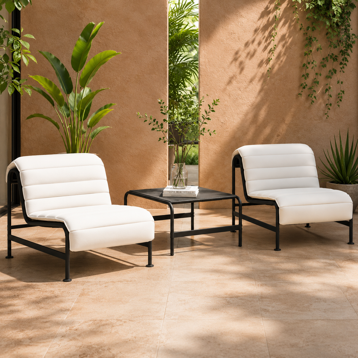 Two white cushioned lounge chairs with black metal frames are placed on a tiled patio, flanking a black square table with a vase of greenery. Lush plants and a beige textured wall complete the setting.