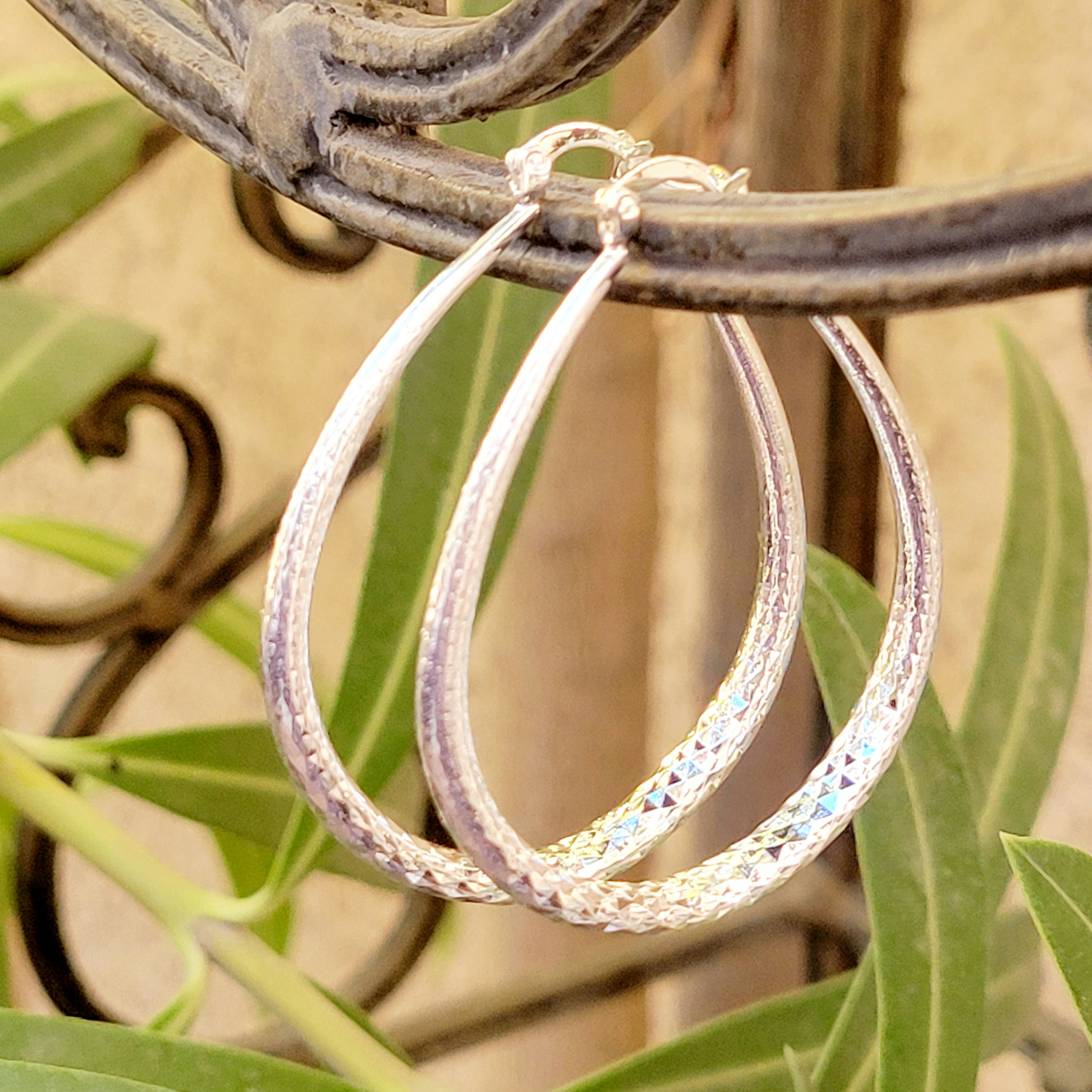 Silver hoop earrings with a textured, diamond-cut design, hanging on a wrought iron railing with greenery in the background.