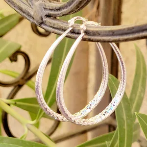 Silver hoop earrings with a textured, diamond-cut design, hanging on a wrought iron railing with greenery in the background.