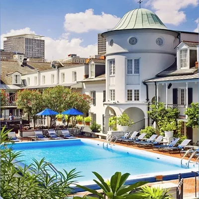 A serene courtyard pool setting surrounded by elegant, white buildings with a distinct circular tower. The area features lounge chairs, blue umbrellas, and lush greenery, set against a backdrop of city buildings.