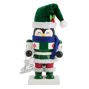 A festive penguin nutcracker dressed in a green Santa hat, a green and red outfit, and a blue scarf is holding a pair of silver skates with a \“Merry Christmas\“ tag.