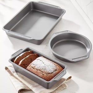 Three non-stick bakeware pans: a rectangular baking dish, a circular cake pan, and a loaf pan filled with sliced powdered bread, all set on a striped kitchen towel.