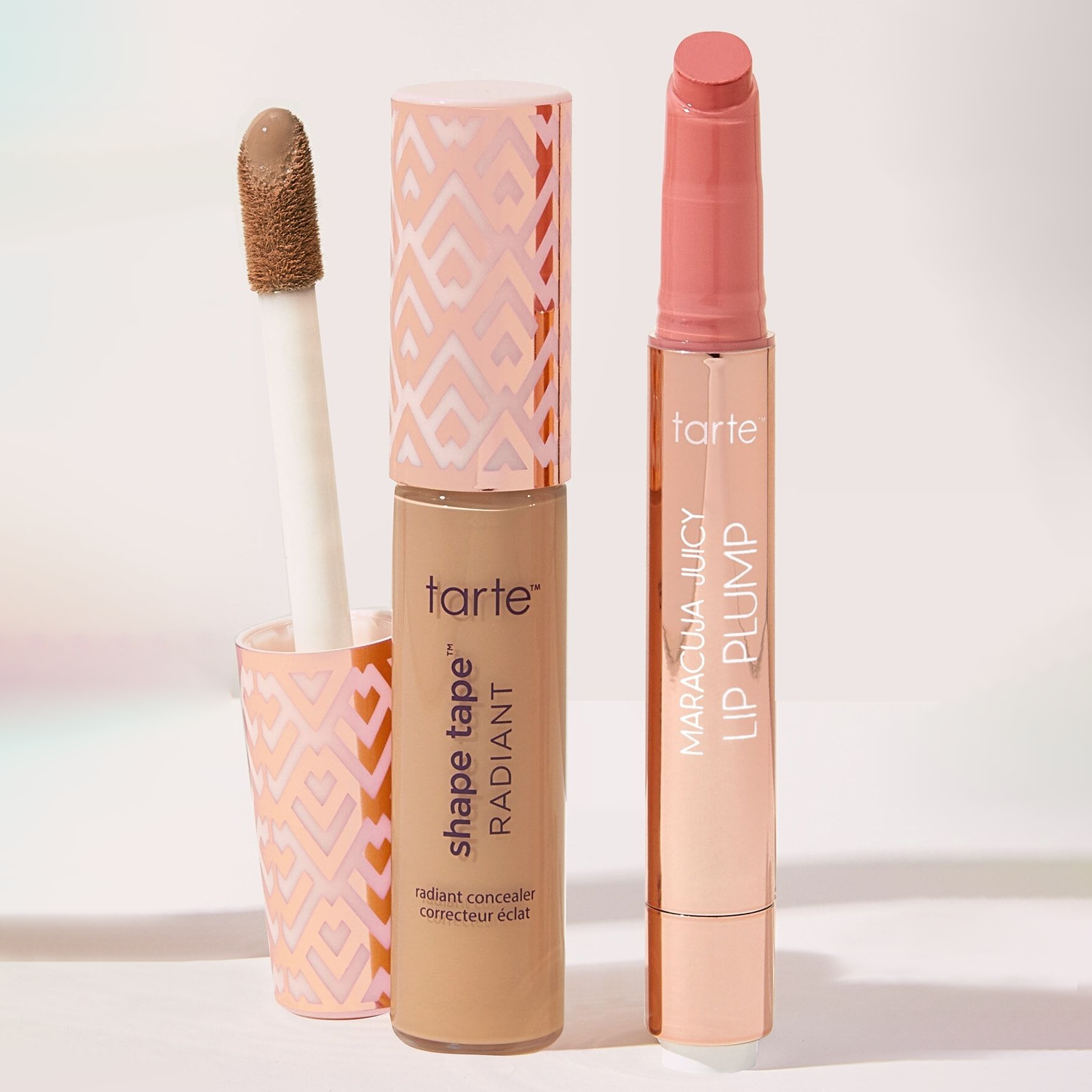 Two Tarte cosmetics items: a Shape Tape Radiant concealer with an open cap and applicator, and a Maracuja Juicy Lip Plump in a twist-up tube.