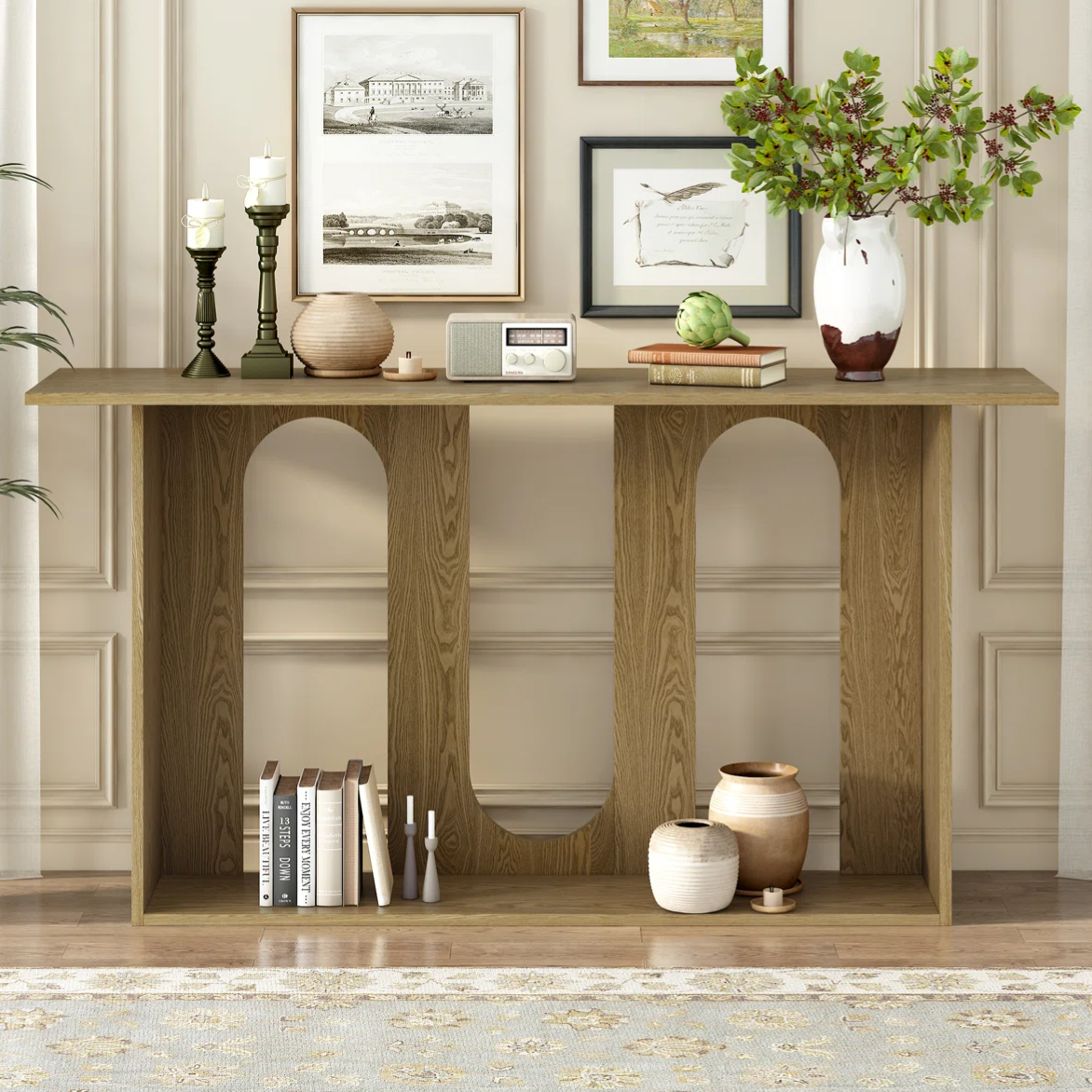 A wooden console table with a geometric design, featuring decorative items like candlesticks, a round vase, books, a small radio, and potted plants. Framed artwork and a soft rug complement the setting.