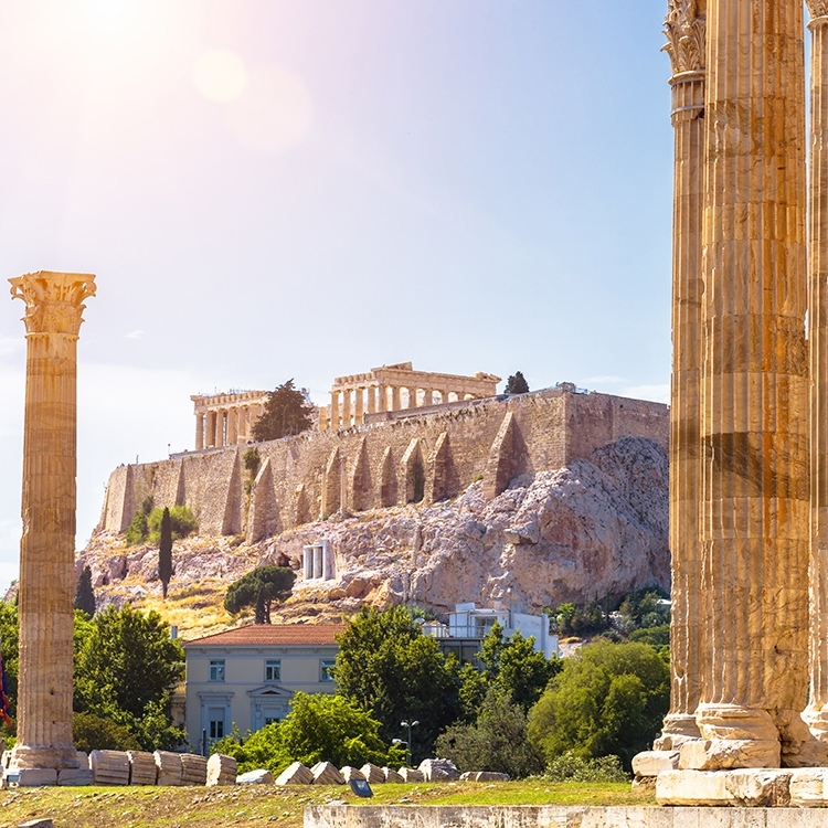 View of the Acropolis in Athens, with ancient Greek columns in the foreground and the Parthenon visible atop the hill, framed by a bright sky and greenery.