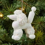 A Christmas tree ornament shaped like a white balloon poodle, covered in sequins and glitter, hangs among the branches.