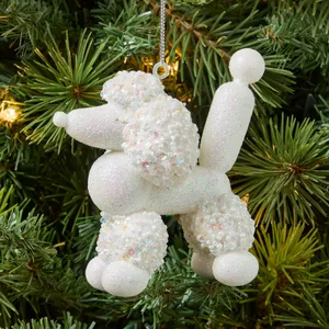 A Christmas tree ornament shaped like a white balloon poodle, covered in sequins and glitter, hangs among the branches.