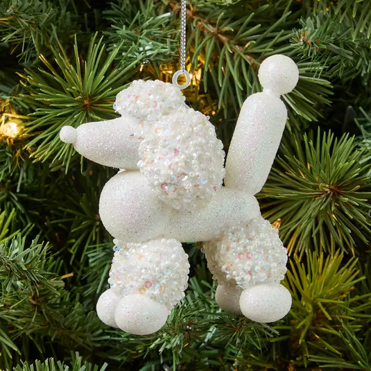 A Christmas tree ornament shaped like a white balloon poodle, covered in sequins and glitter, hangs among the branches.