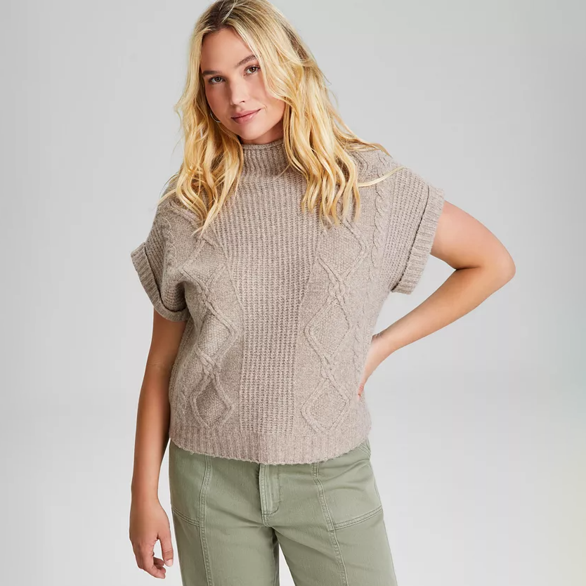 A beige, cable-knit sweater vest with a funnel neck and ribbed trim, featuring a sleeveless design and a cropped length.