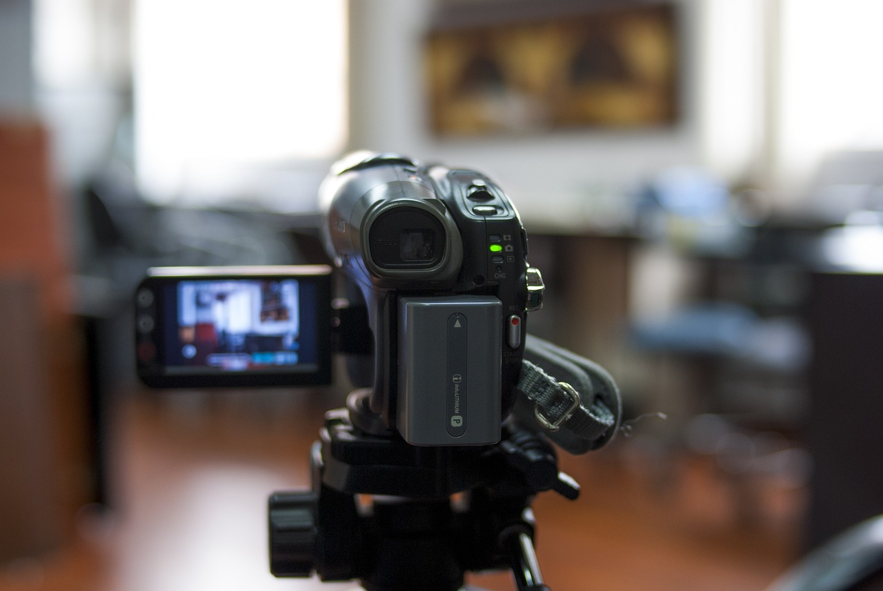 A digital camcorder on a tripod, facing a blurry indoor background.