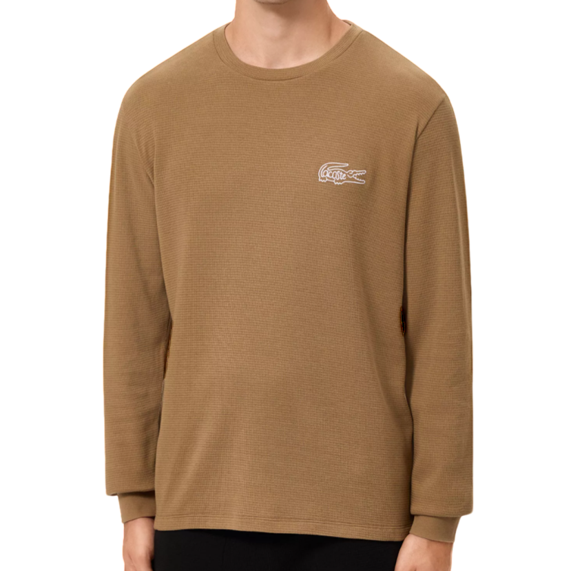 A man is wearing a brown Lacoste long-sleeve crew neck T-shirt with a white logo on the chest. The shirt is paired with black pants.