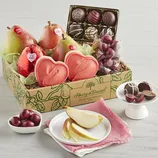 A gift box from Harry & David featuring pears, heart-shaped cookies, chocolates, and cherries, displayed with a small plate of pear slices and chocolates on a stand.