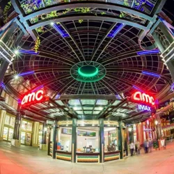 A brightly lit AMC and IMAX theater entrance with colorful lighting and a modern design. The scene includes a ticket booth and a few people walking by.
