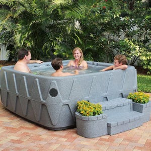 A square hot tub with three people enjoying it, set in a garden with surrounding tropical plants and flowers in planters.