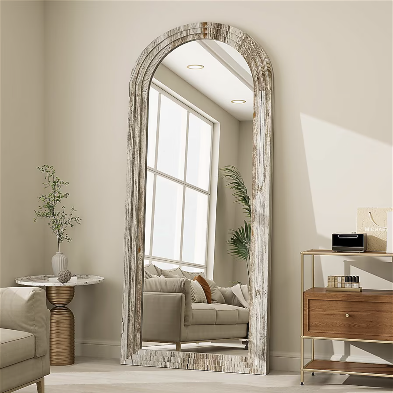 An arched floor mirror with a distressed wood frame stands in a living room setting, reflecting a sofa, a marble-topped side table, and a wooden console with decor items.
