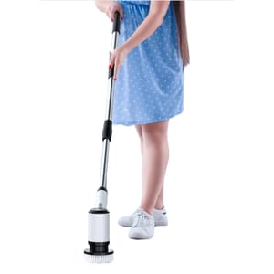 A person is using an electric scrub brush with an extendable handle for cleaning, wearing a blue polka dot dress and white sneakers.