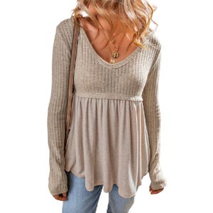 A beige long-sleeve babydoll top with a ribbed, fitted bodice and a flowy, gathered lower section. It features a V-neckline and is paired with blue jeans and layered gold necklaces.