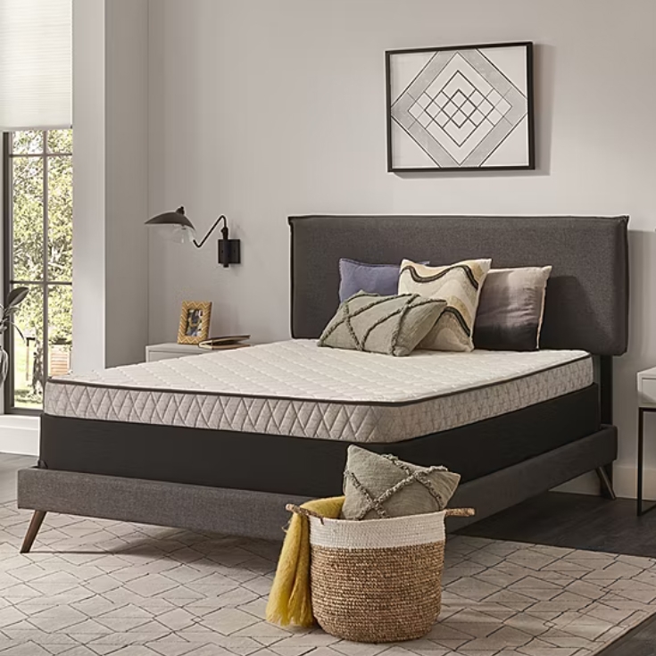 Queen-size mattress with a plush top and medium firmness, featuring a quilted pattern. It rests on a stylish gray upholstered bed frame with angled legs, complemented by decorative pillows and a woven basket with a throw.