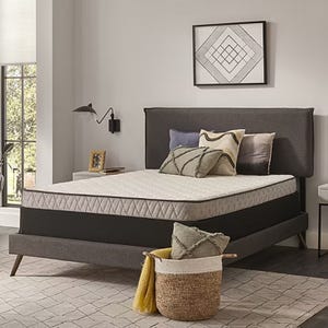 Queen-size mattress with a plush top and medium firmness, featuring a quilted pattern. It rests on a stylish gray upholstered bed frame with angled legs, complemented by decorative pillows and a woven basket with a throw.