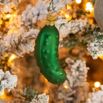 A green pickle-shaped ornament hangs on a snow-frosted Christmas tree, illuminated by warm lights.