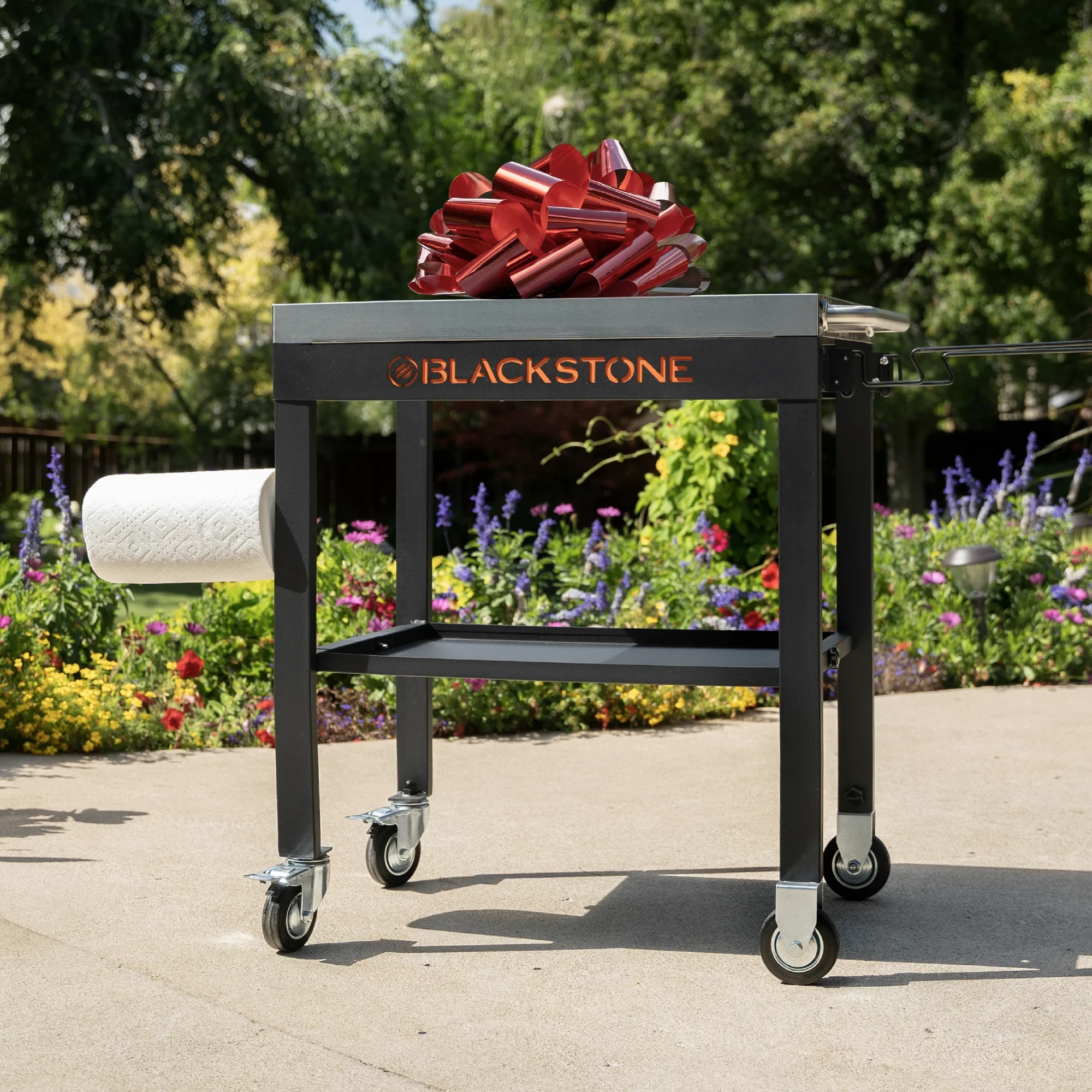 A Blackstone outdoor cooking cart with a large red bow on top and a roll of paper towels hanging on the side, positioned on a pathway with a garden in the background.