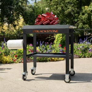 A Blackstone outdoor cooking cart with a large red bow on top and a roll of paper towels hanging on the side, positioned on a pathway with a garden in the background.