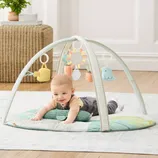 A baby is on a play mat featuring an arched mobile with hanging toys including a watering can, sun, and cloud. The setup rests on a wooden floor in a brightly lit room.