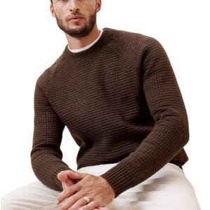 A man is wearing a brown, cozy textured sweater with a ribbed crew neck, cuffs, and hem, paired with white pants. The sweater has a subtle grid pattern and appears comfortable and casual.