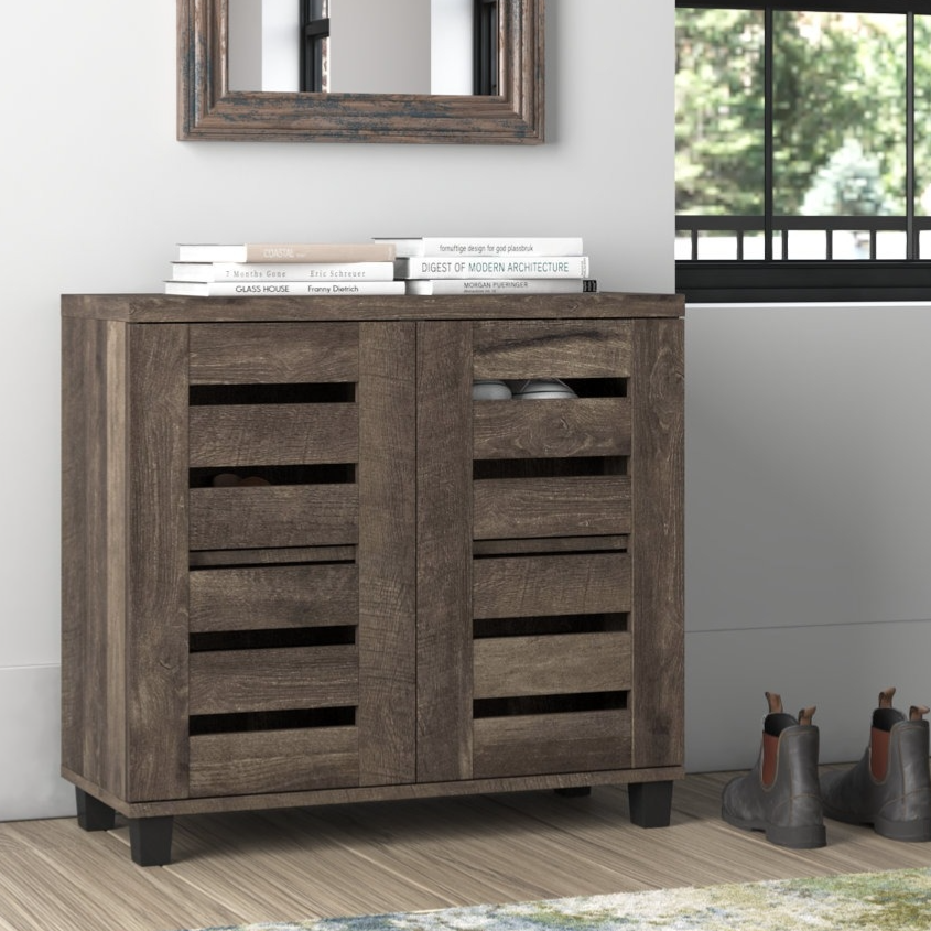 Wooden shoe cabinet with slatted doors, approximately 31.5'' H. Topped with books, it stands beside a window and some boots, with a decorative mirror above.