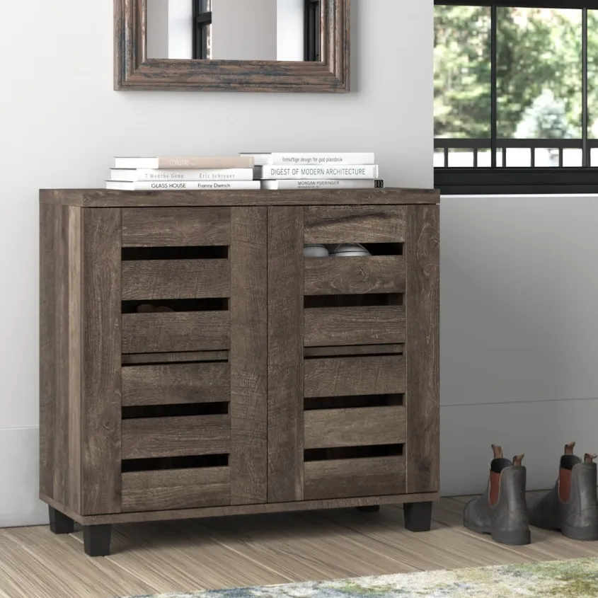 Wooden shoe cabinet with slatted doors, approximately 31.5'' H. Topped with books, it stands beside a window and some boots, with a decorative mirror above.