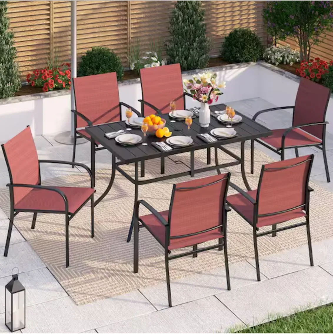Outdoor dining set with black table and six red cushioned chairs, set on a light-colored rug. The table is adorned with plates, glasses, a vase with flowers, and a bowl of fruits. The background features potted plants and a wooden fence.