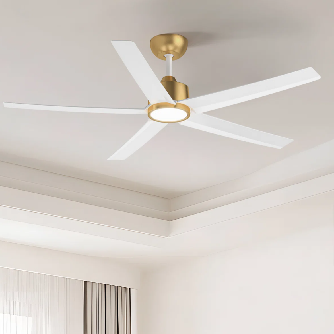 A modern ceiling fan with a gold and white finish, featuring five blades and an integrated light fixture, installed in a well-lit room with a tray ceiling and curtains.