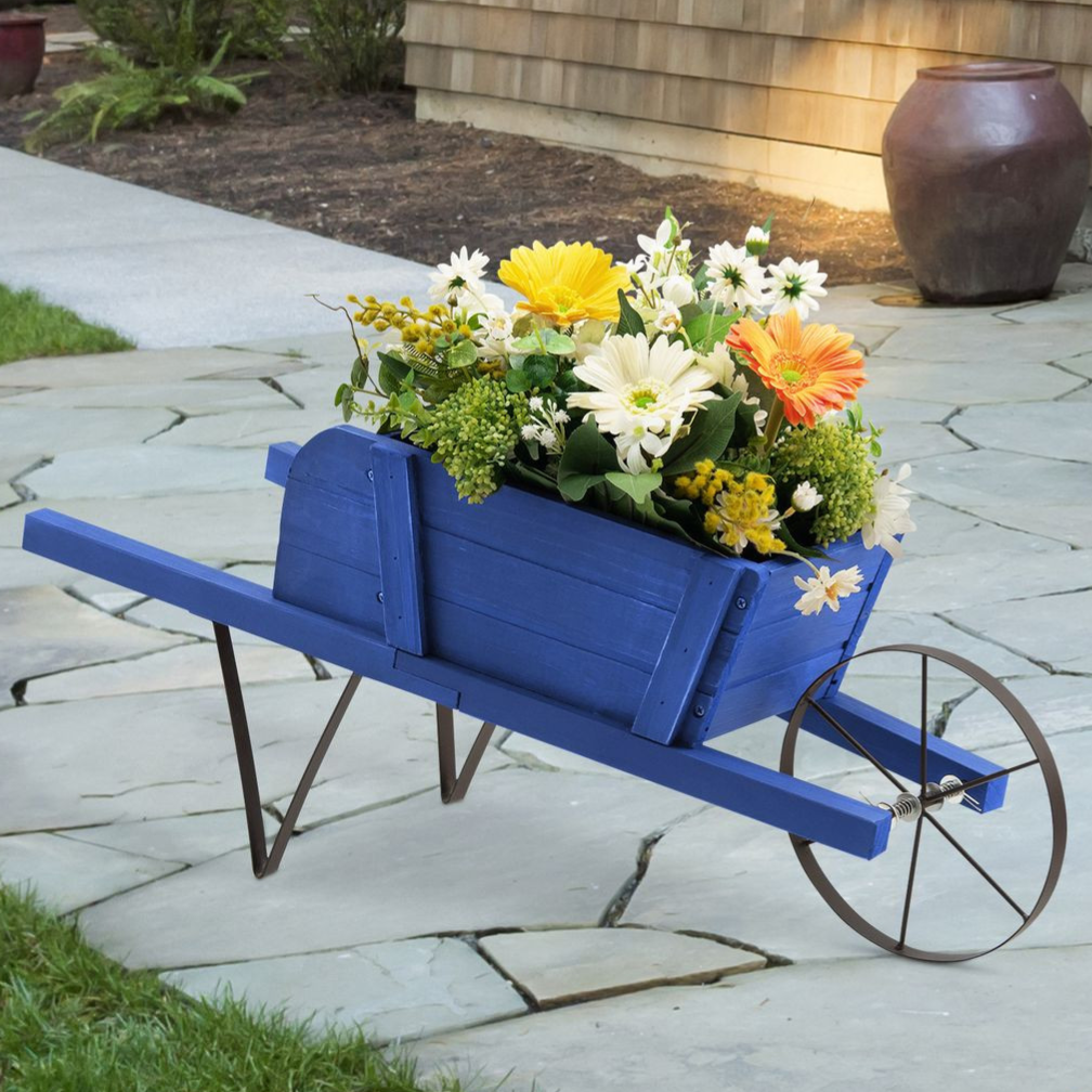 A blue wooden wheelbarrow planter filled with vibrant yellow, white, and orange flowers, set on a paved patio with grass and a large ceramic pot in the background.