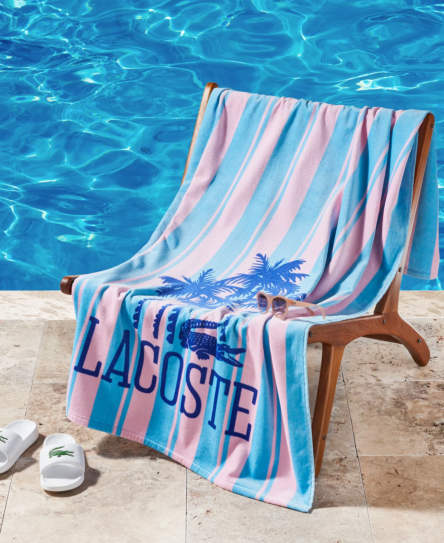 Poolside setup with a striped Lacoste towel featuring palm trees and a pair of sunglasses on a wooden chair, next to a clear blue pool. White Lacoste slides are placed on the stone deck.