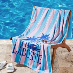 Poolside setup with a striped Lacoste towel featuring palm trees and a pair of sunglasses on a wooden chair, next to a clear blue pool. White Lacoste slides are placed on the stone deck.