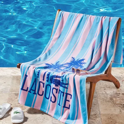 Poolside setup with a striped Lacoste towel featuring palm trees and a pair of sunglasses on a wooden chair, next to a clear blue pool. White Lacoste slides are placed on the stone deck.